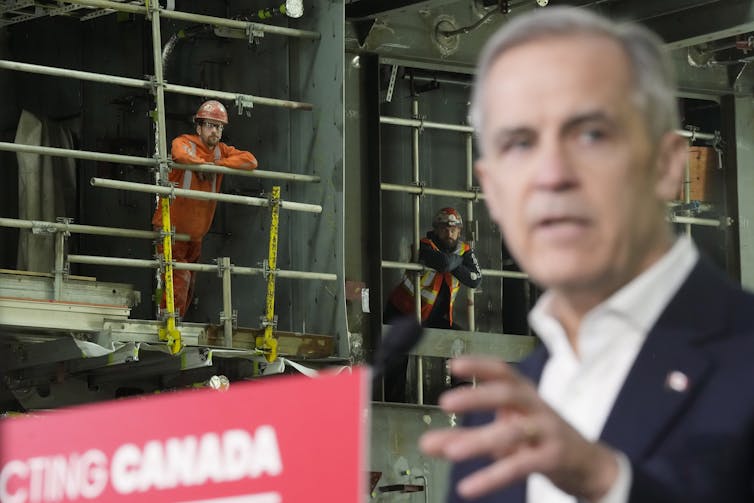 A man in an orange safety suit is in focus as listens from behind a blurred man with short grey hair.