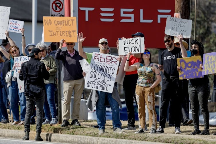 Protesters with signs in front of large Tesla logo
