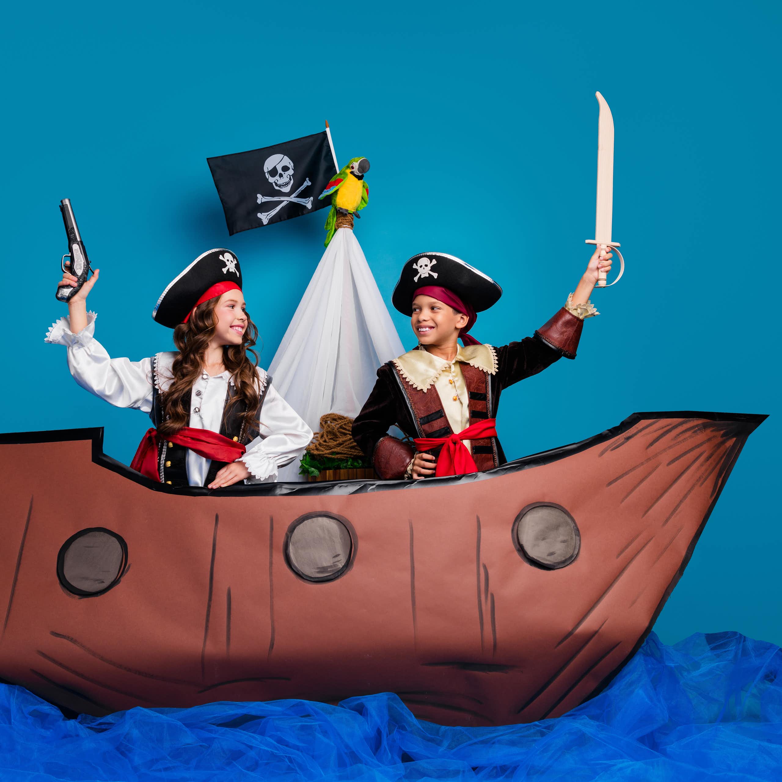 Two children, a boy and a girl, are dressed in pirate costumes on a fake boat in a staged setting.