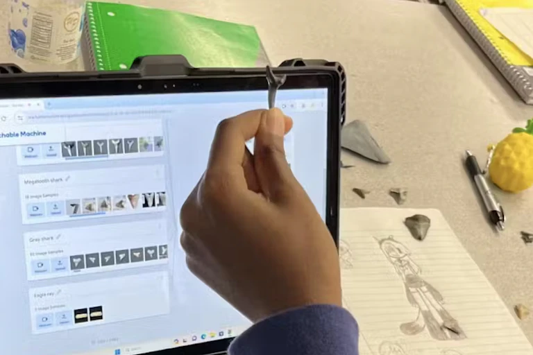 A hand holds up a shark's tooth in front of an iPad with other teeth visible on a screen. Other shark teeth, paper and pencil are on a desk nearby.