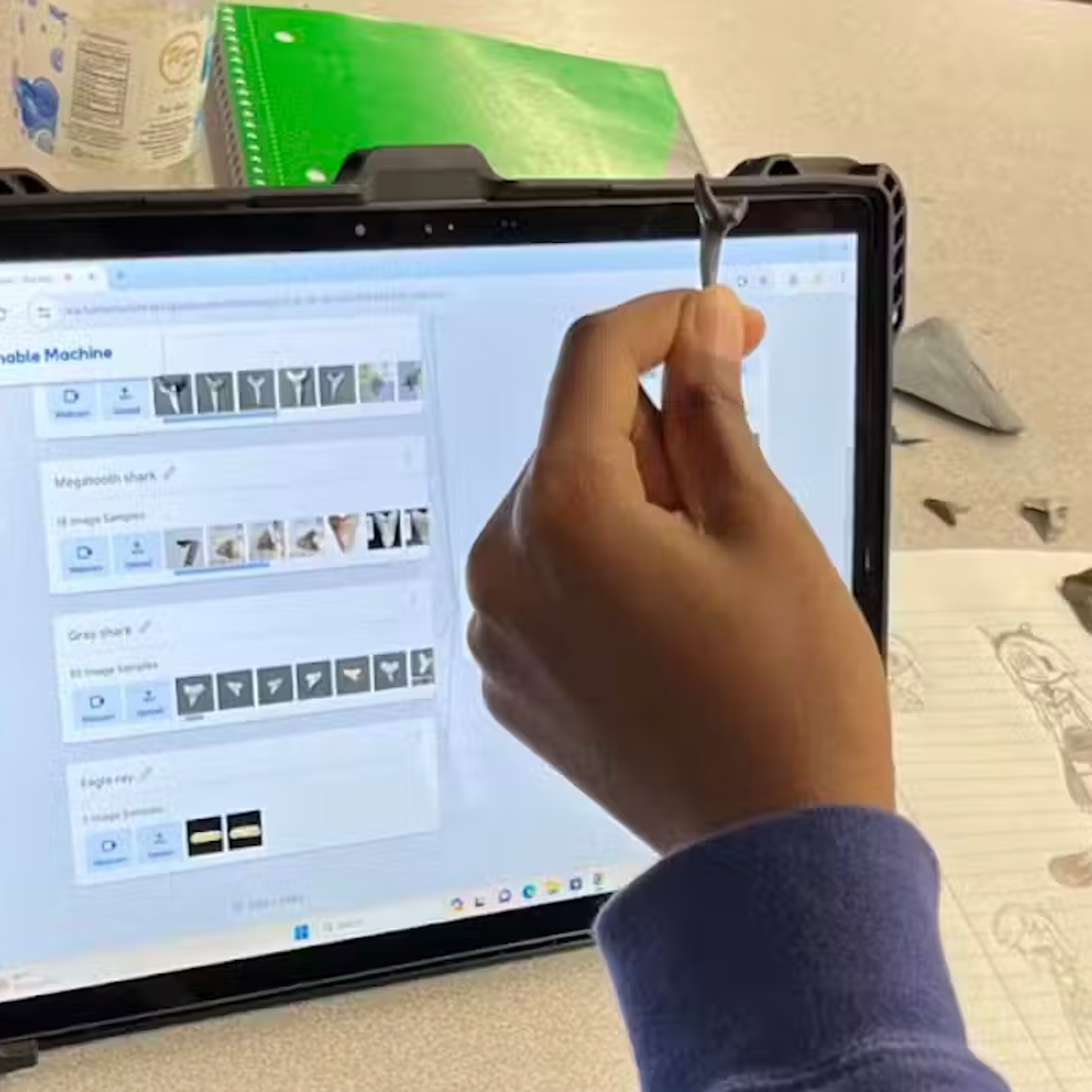 A hand holds up a shark's tooth in front of an iPad with other teeth visible on a screen. Other shark teeth, paper and pencil are on a desk nearby.