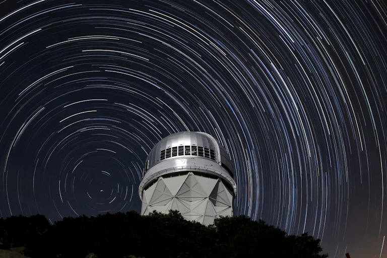 An observatory on a hill, with streaks in the sky in the background representing stars moving over time.