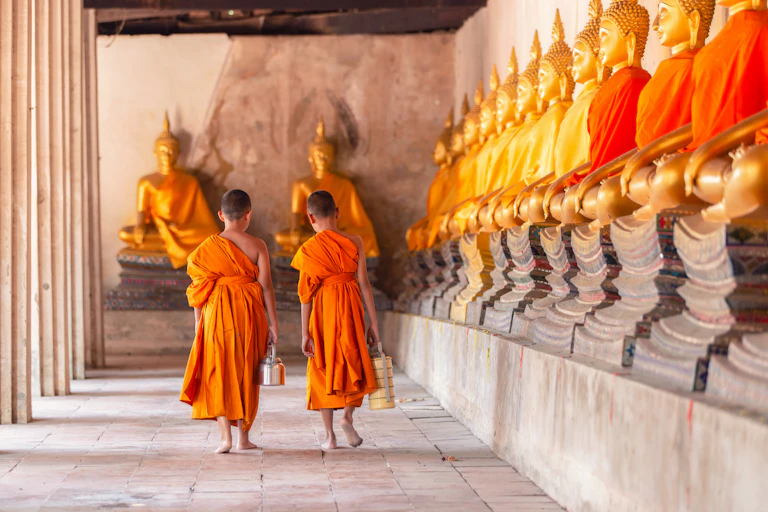 Two monks in saffron robes stand before Buddha icons arranged along the front and side walls of a temple.