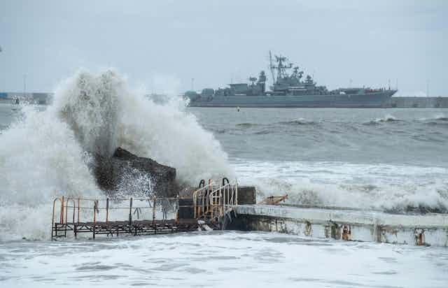 A large warship is seen at sea behind a big wave crashing on some rocks