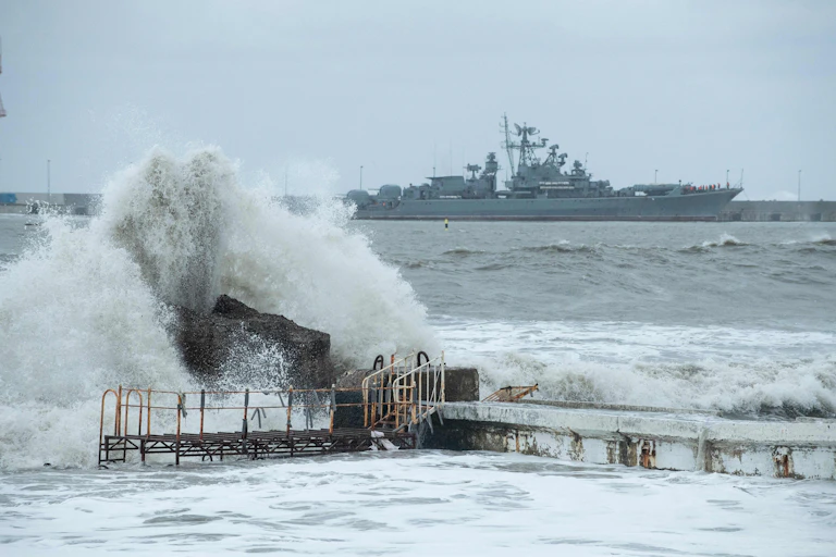 A large warship is seen at sea behind a big wave crashing on some rocks