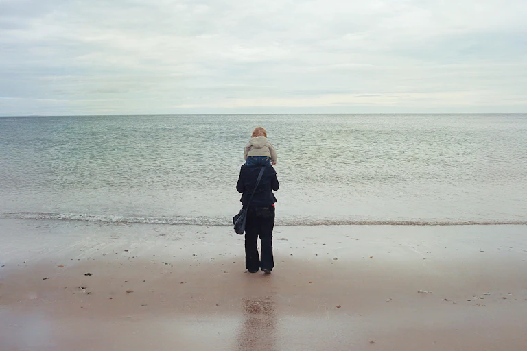 A mother and son at the beach, he sits on her shoulders as they look out to sea.