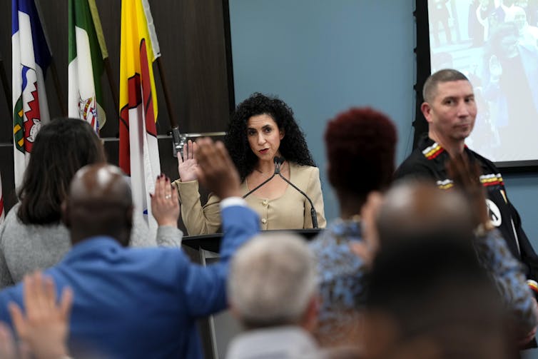 Una mujer con cabello negro largo y rizado habla detrás del podio a un montón de personas con un arma elevada