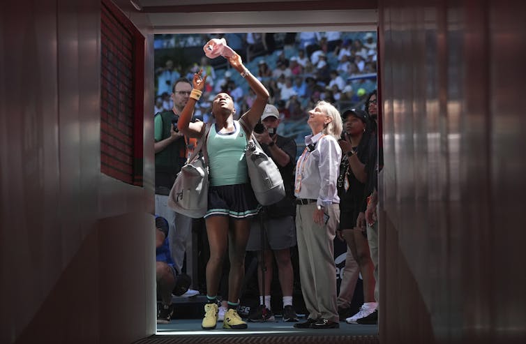 A Black woman in a tennis uniform passes a baseball hat upwards, presumably to fan sitting in the stands of a stadium off camera