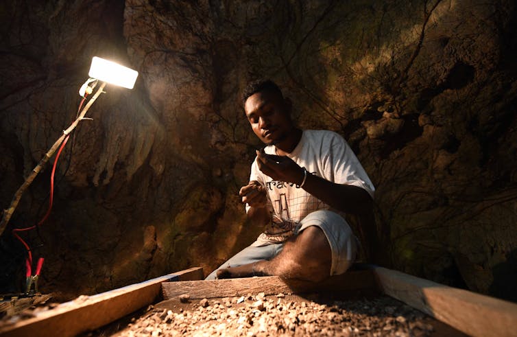 A man working under the light of a lamp, examining excavated artefacts at Mololo Cave.