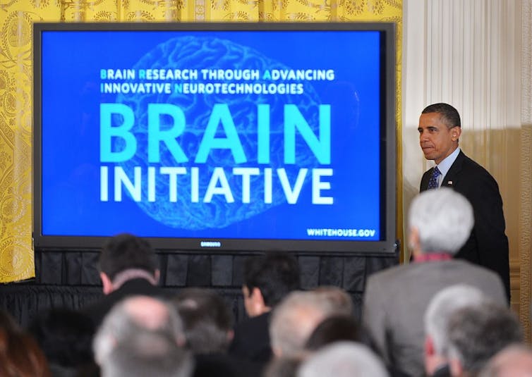 Former president Barack Obama standing beside a screen reading 'BRAIN INITIATIVE' at a press conference