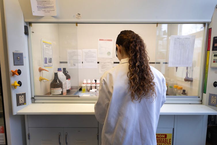 Person in white coat standing before a fume hood, back to camera