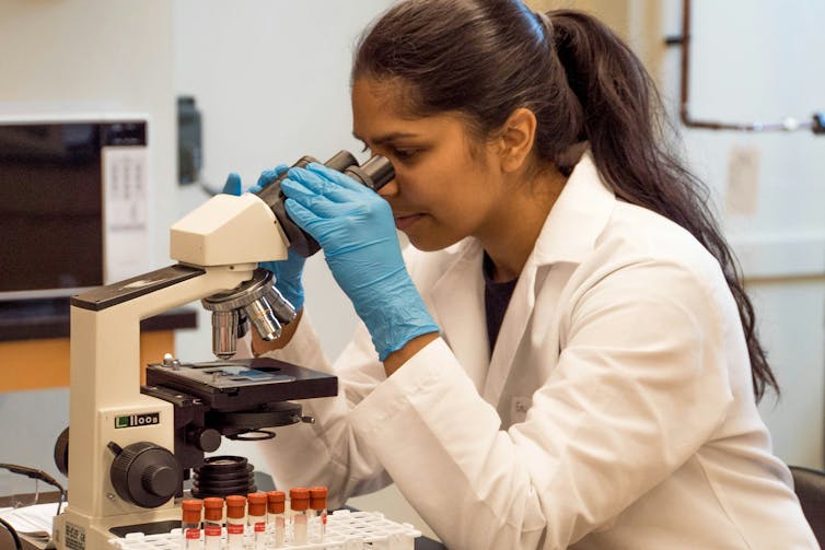 Woman with brown skin wearing a lab coat looking through a microscope.