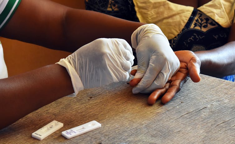 Gloved hands perform a finger prick blood test on another person's hand.