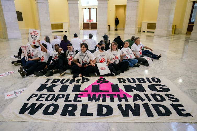 People sitting on the floor with a large sign which reads TRUMP & RUBIO KILLING PEOPLE WITH AIDS WORLDWIDE.