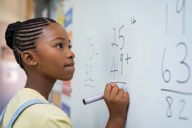 Schoolgirl solving addition sum on white board with marker pen.