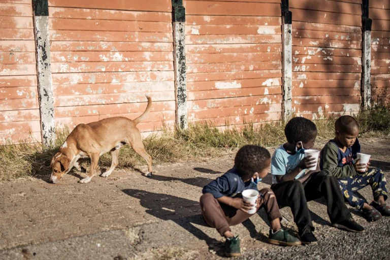 Three young children sit on a pavement, holding disposable cups, while a dog sniffs the ground behind them