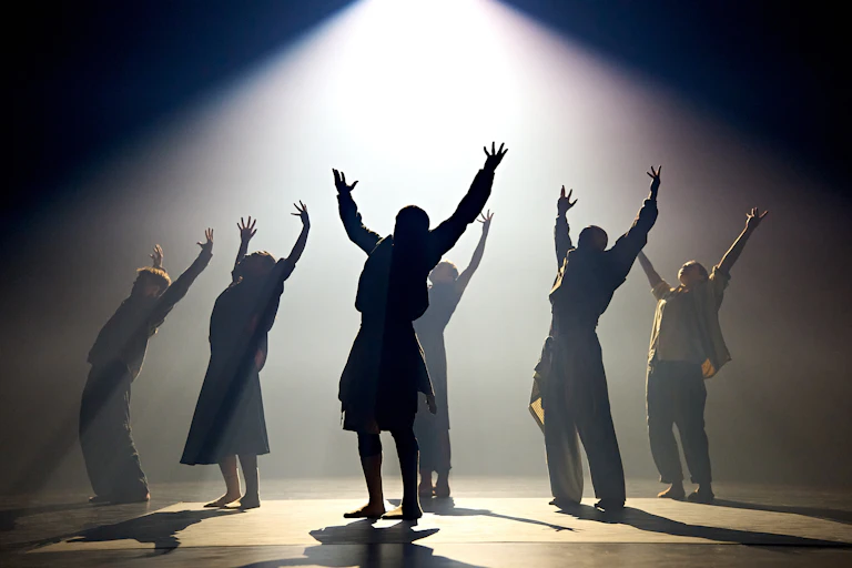 Shadowy figures on stage their arms stretch out wide downlit by a spotlight.