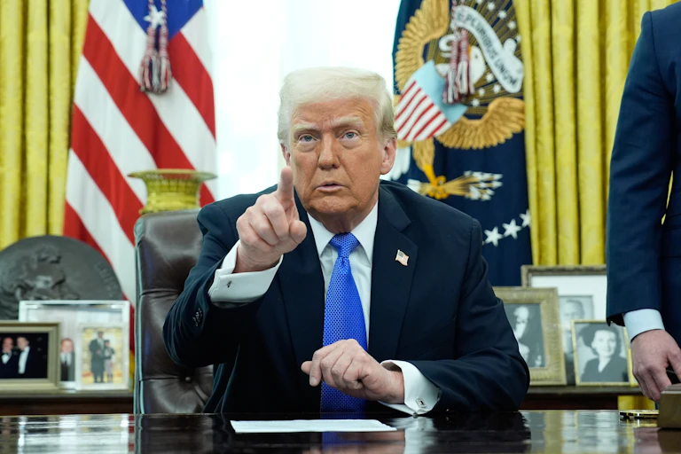 Donald Trump sits behind the Resolute Desk in the Oval Office and pints at the camera, March 21 2025.