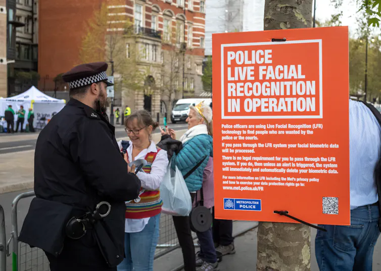 Next to a blocked-off road for an event, a police officer stands by a sign that alerts the public to a police live facial recognition operation in progress.