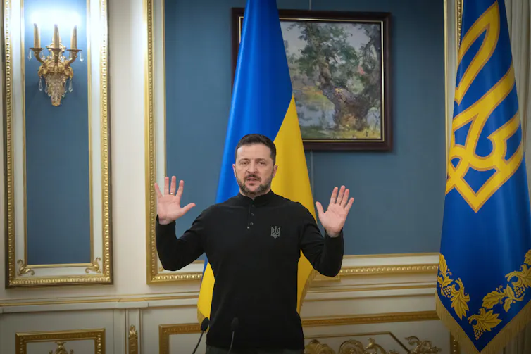 A man in dark clothing with dark hair and a beard with both hands raised as he speaks in front of a Ukrainian flag.