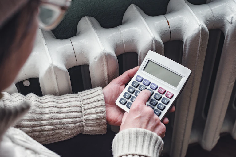 An elderly woman uses a calculator next to a radiator.
