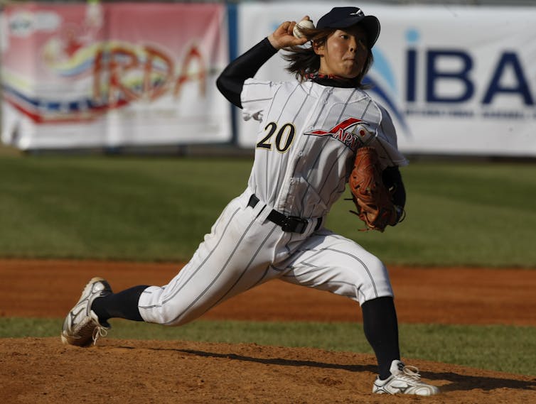 Young Japanese woman wearing a baseball uniform throws a pitch from a mound.
