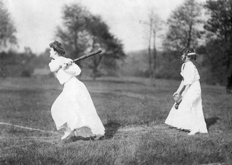 Black and white photo of two women wearing white dresses playing baseball in a grassy field.