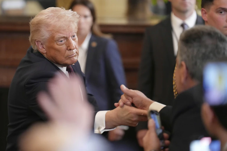An older man with orange-tinged grey hair and heavy facial bronzer shakes hands with someone whose back is to the camera.