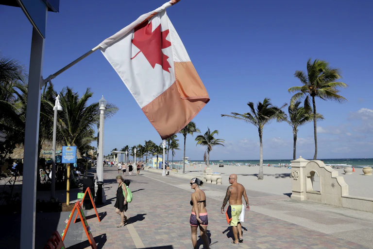 A Canadian flag flies near a sandy beach as people walk along a boardwalk.
