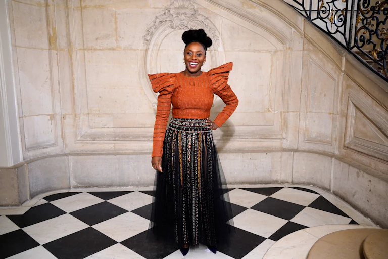 An African woman with an elaborate hairstyle smiles broadly as she poses near a vintage staircase, wearing a black skirt and an orange blouse with winged shoulders.