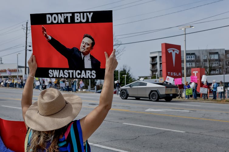 A woman holds up a sign in front of a Tesla dealership reading 'don't buy a swasticar'.