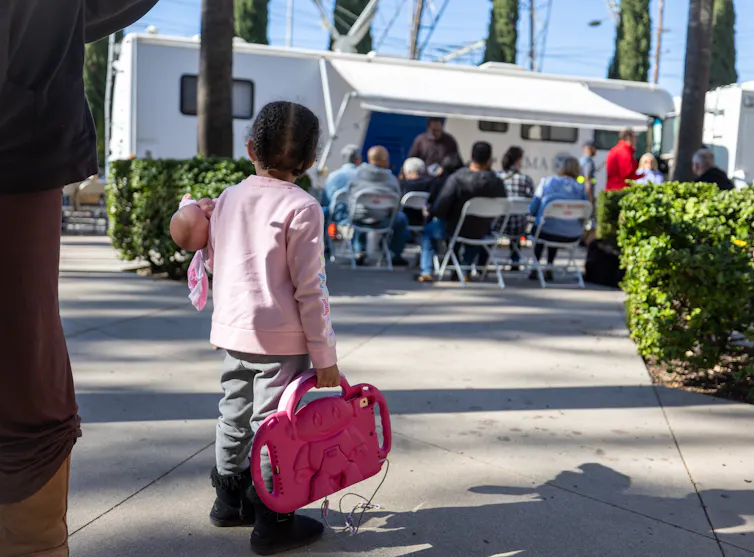 Little girl holding iPad watches adults meeting outside a trailer.