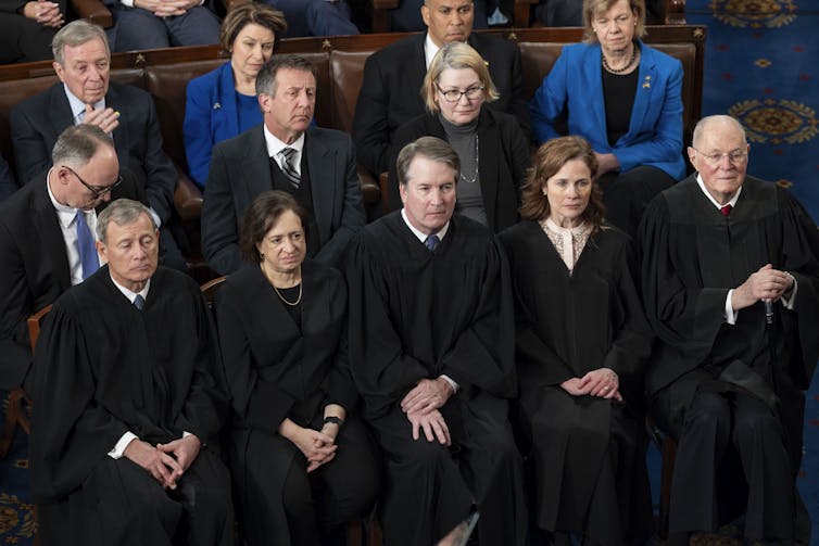 A row of seated men and women in black robes, with suited men and women sat behind them.
