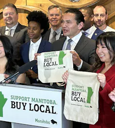 Derrière un podium, des personnes brandissent des sacs en tissu sur lesquels on peut lire « buy local » (achetez local).’