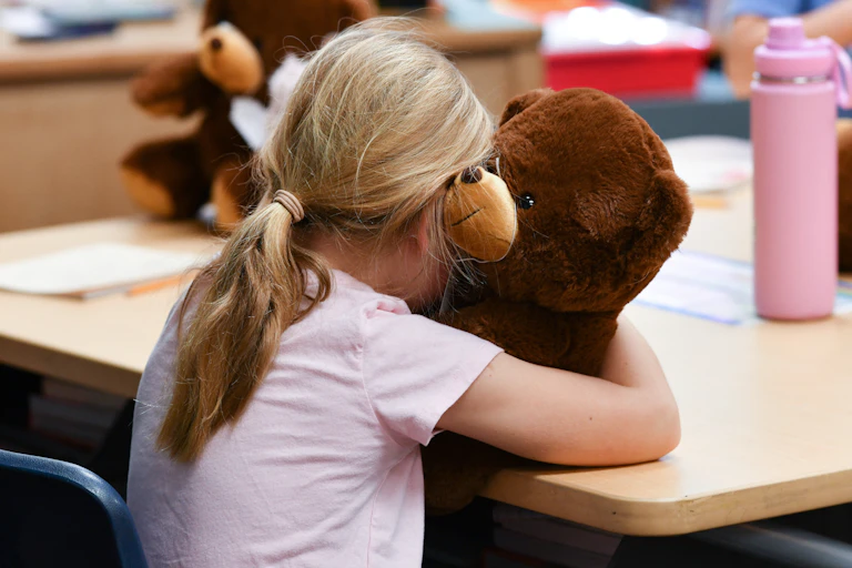 Girl clutches teddy bear at school desk