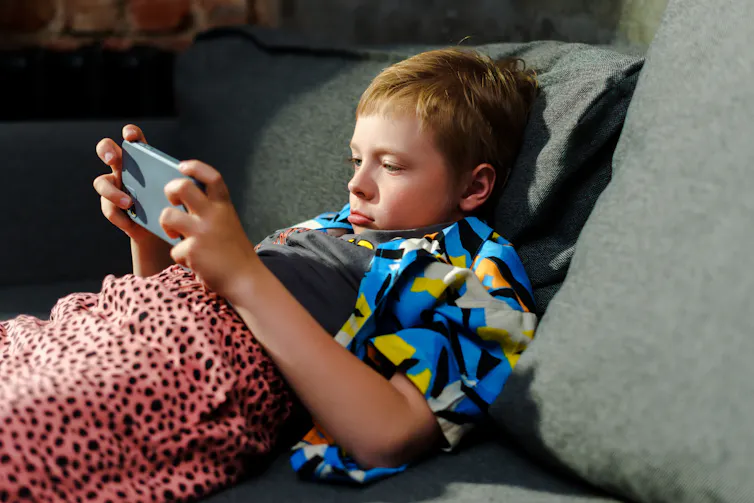 A young boy pouts, while lying on the couch with a phone and a blanket.