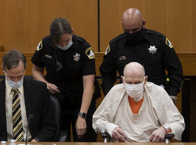 a bald man in prison uniform in a wheelchair. two police officers are standing behind him and a man in a suit sits next to him. all are wearing surgical face masks.
