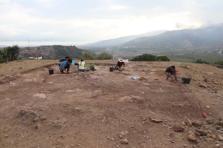Four people kneel as they excavate an earthy site, mountains in the distance.