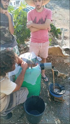 Two men stand and watch a third who has his hand in a green bucket.