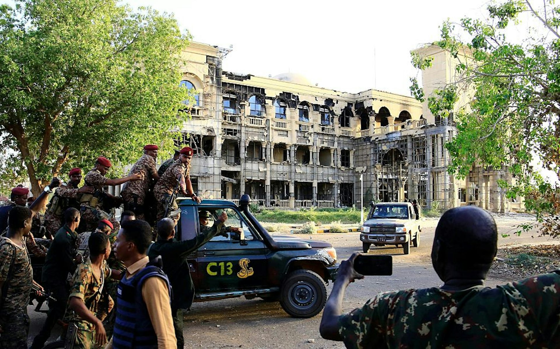 Men in military fatigues aboard a pickup vehicle written C13 drive near a three-storey building that has a damaged exterior.