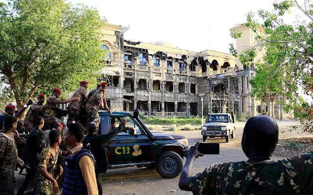 Men in military fatigues aboard a pickup vehicle written C13 drive near a three-storey building that has a damaged exterior.