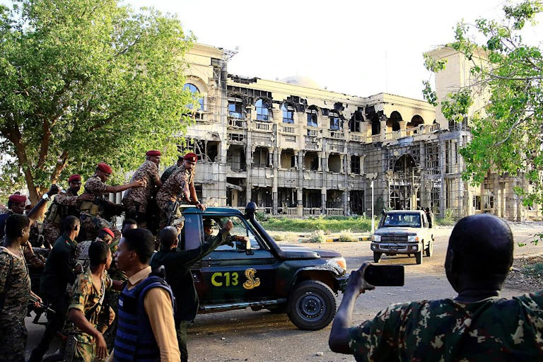 Men in military fatigues aboard a pickup vehicle written C13 drive near a three-storey building that has a damaged exterior.