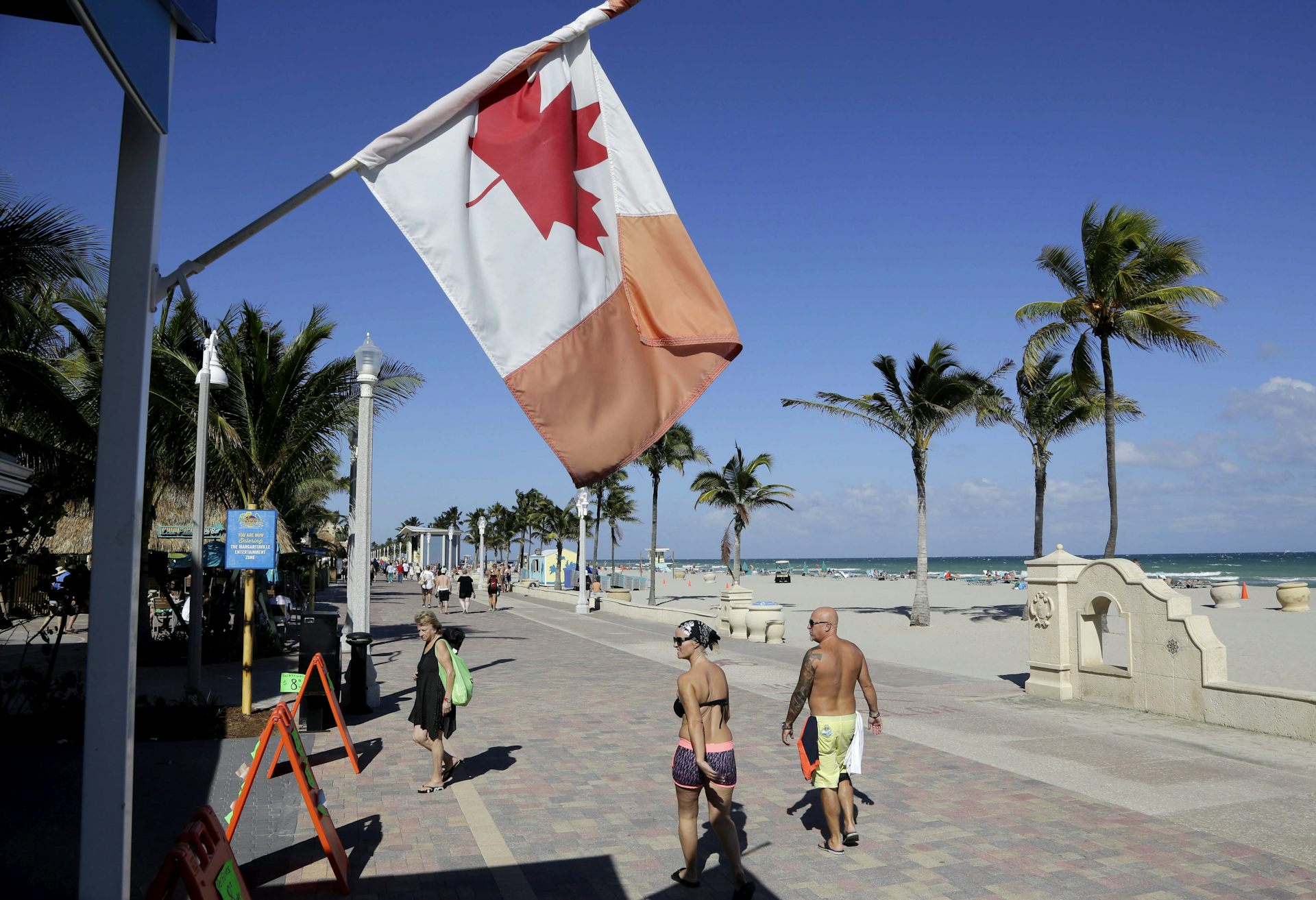 Un drapeau canadien flotte près d'une plage de sable, tandis que des gens marchent le long d'une promenade.