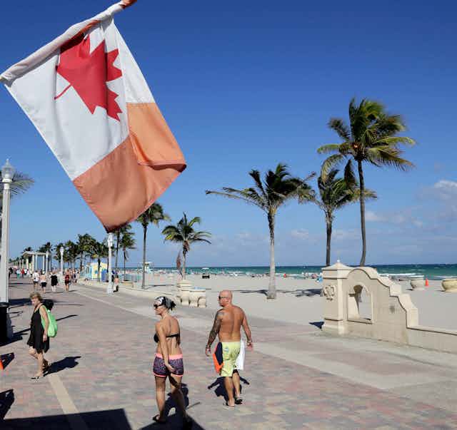 Un drapeau canadien flotte près d'une plage de sable, tandis que des gens marchent le long d'une promenade.