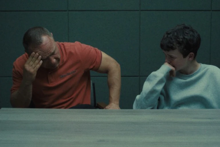 A man holds his head while a youth leans on his fist in a grim cinderblock room with dim lighting.