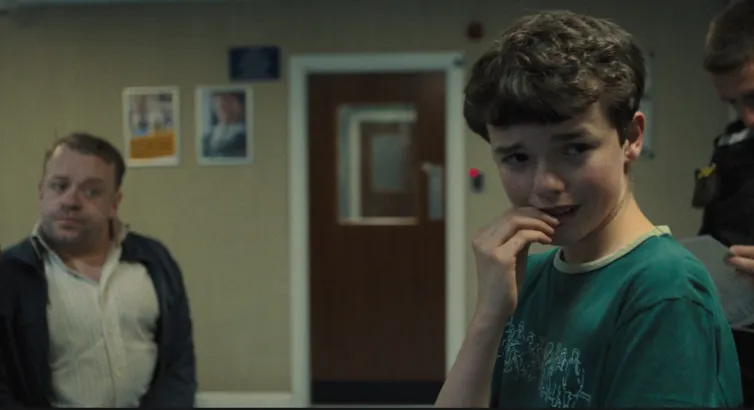 A boy looking anguished in a dark room with an adult watching him.