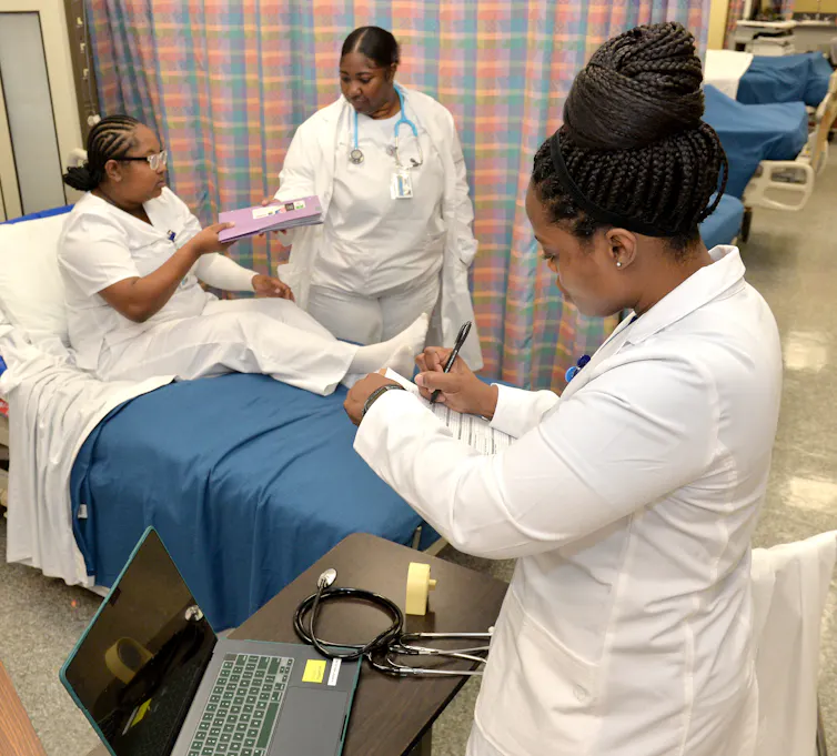 Three women dressed in white scrubs and lab coats in a hospital room