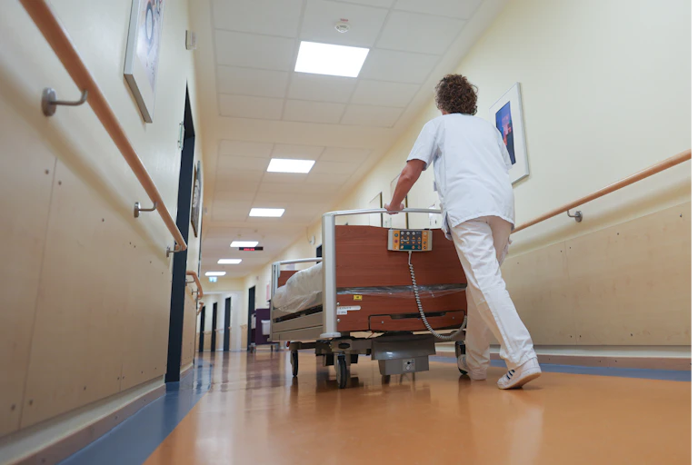 Nurse in white scrubs seen pushing hospital bed down corridor.