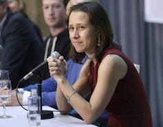 A woman with dark hair in a red sleeveless with a microphone on a table in front of her.