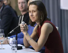 A woman with dark hair in a red sleeveless with a microphone on a table in front of her.